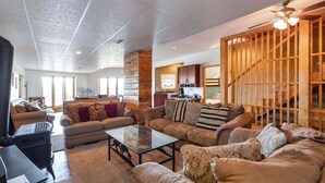 A spacious living room with a fireplace, sofas, and a coffee table. A kitchen is visible in the background, and there is a wooden staircase. - 5BR Table Rock Lake Getaway |Hot Tub, Pool Games (Blue eye)