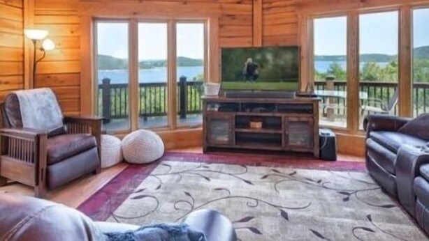 A cozy cabin interior with a large TV, wooden walls, and a scenic lake view. - 5BR Table Rock Lake Getaway |Hot Tub, Pool Games (Blue eye)