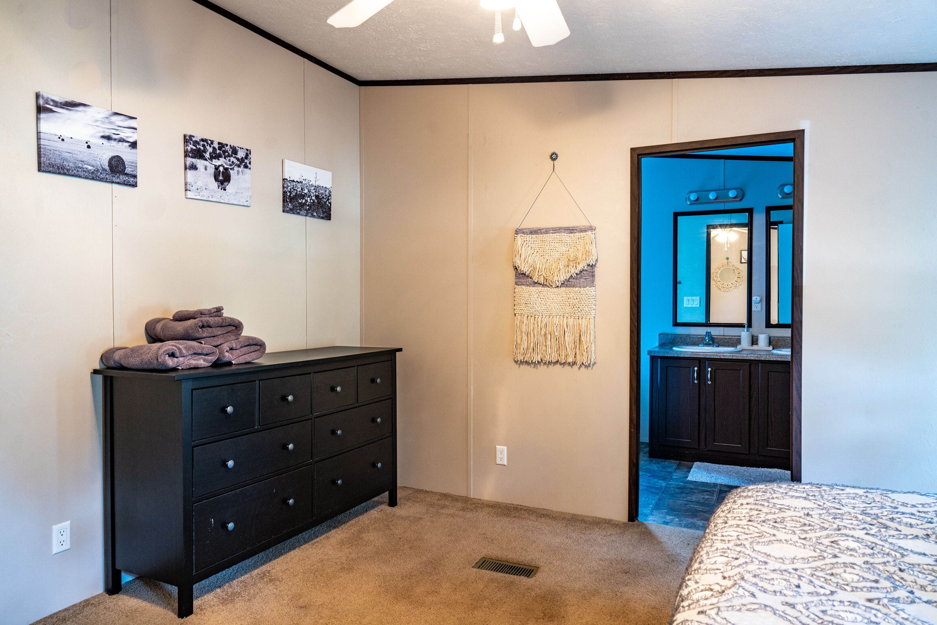 Een slaapkamer met een bed, een dressoir, een macramé wanddecoratie en een deur naar een badkamer.