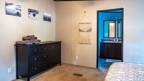 A bedroom with a bed, a dresser, a wall-mounted macrame piece, and a door leading to a bathroom. - $0 Cleaning - Best for Groups and Families (Bastrop)