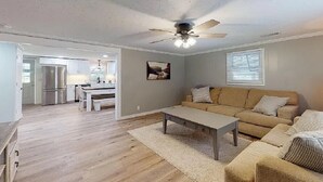 A spacious living room with a beige sofa, a coffee table, and a ceiling fan. A kitchen is visible in the background.