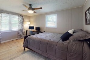 Ein Schlafzimmer mit einem Bett, einem Schreibtisch mit Computer, einem Deckenventilator und zwei Fenster mit Jalousien.
