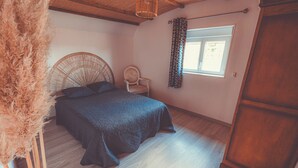 A bedroom with a bed, a wicker chair, a window with curtains, and a wooden wardrobe. - House in the heart of the Périgord Vert. (Payzac)