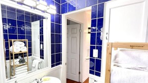 A bathroom with a white sink, mirror, and blue-tiled walls. There is a wooden shelf with towels and a door leading to another room.