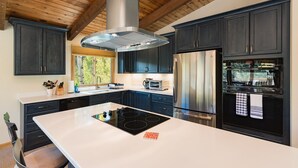 A modern kitchen with dark cabinetry, a central island with an induction cooktop, a stainless steel refrigerator, and a double oven. - Cabin on Columbine Lake - Large deck - Hot Tub/Views  - 1 mile to Purgatory (Durango)