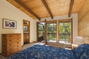 Een slaapkamer met een houten plafond, een bed met een blauw bloemenbedekking, een dressoir, een raam met uitzicht op bomen en een deur naar een balkon.