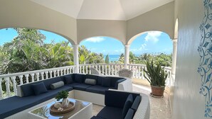 Un área de estar al aire libre techada con vistas al océano, columnas blancas y una planta en maceta.