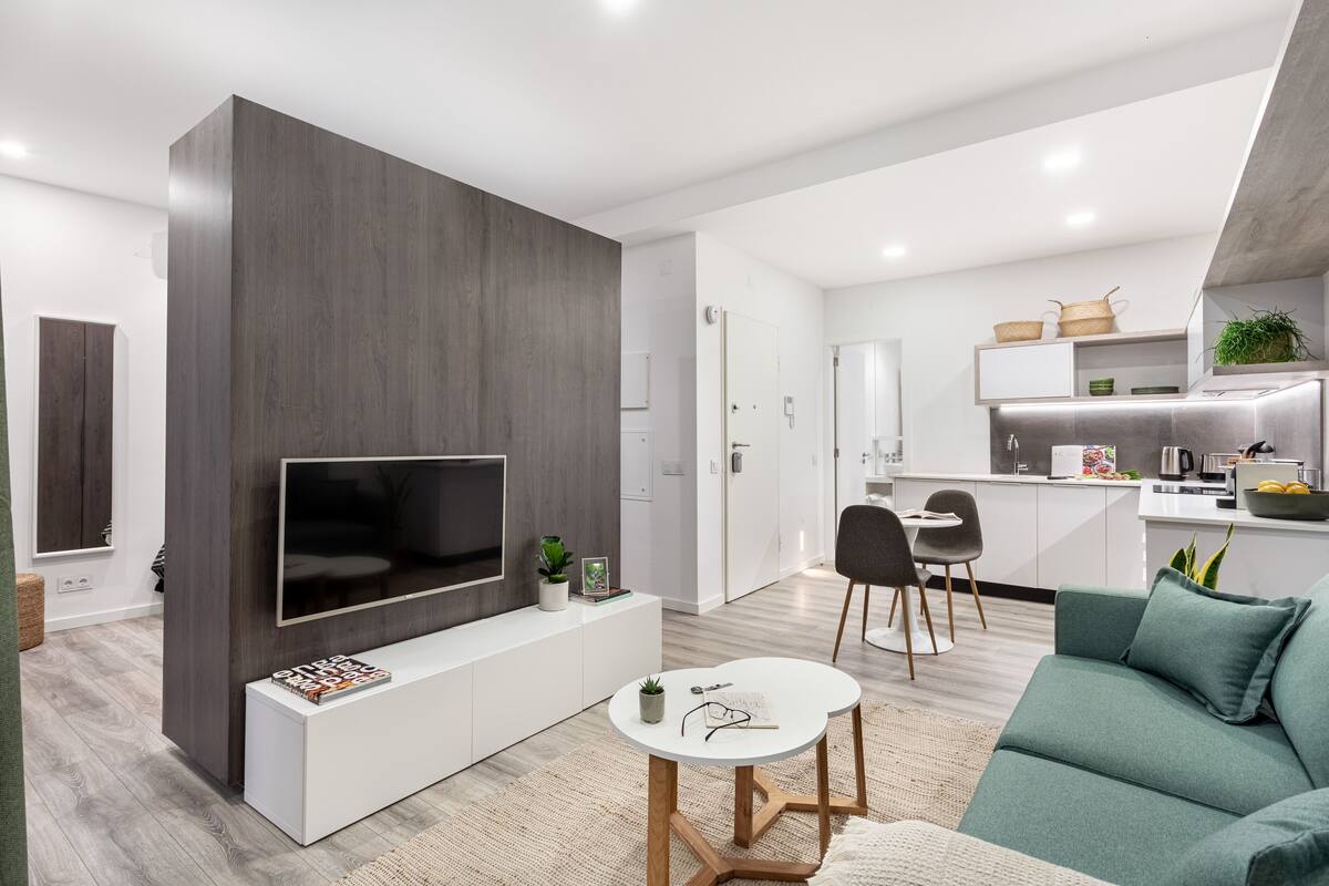 Un salon moderne avec un téléviseur à écran plat, une console blanche, un canapé vert et un coin cuisine à l’arrière-plan.