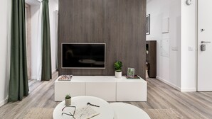 A modern living room with a flat-screen TV mounted on a wooden panel wall, a white entertainment unit with a plant and picture frame, and a round coffee table with reading materials. - Studios Terra & Mar (Lisbon)