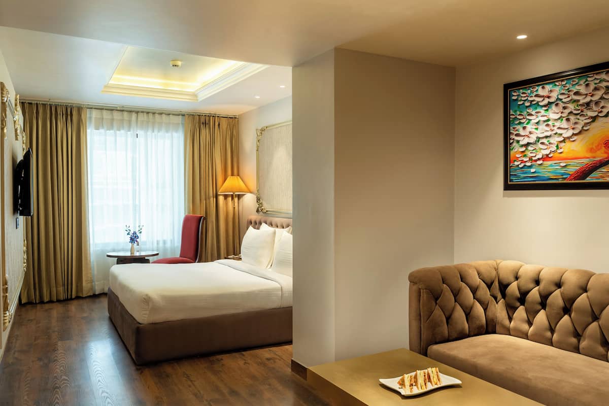 A hotel room with a bed, a sofa, a table with a tray of food, a lamp, and a painting on the wall.
