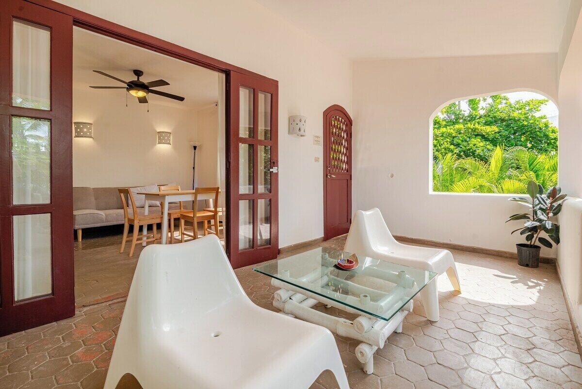 Eine Terrasse mit Glastisch, weißen Stühlen, Deckenventilator und Blick ins Grüne durch ein Fenster.