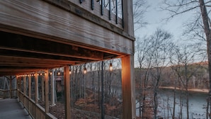A wooden deck with string lights, overlooking a lake and forested area. - Private Dock | Hot Tub | Kayaks | GameRoom | EV (Dawsonville)