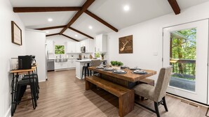 A modern kitchen with white cabinets, a wooden dining table, and a bench. There is a large window with a view of greenery. - Private Dock | Hot Tub | Kayaks | GameRoom | EV (Dawsonville)