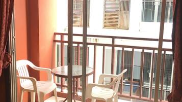 A balcony with two white plastic chairs and a small round table, a glass door, and a view of buildings with shutters.