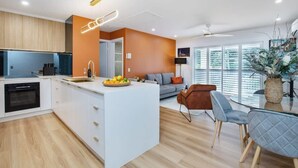 A modern kitchen with white cabinets, a central island with a fruit bowl, and a dining area with a glass table and chairs. - Top Floor in Burleigh: Pool Parking Walk To Beach (Gold Coast)