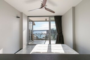 A bedroom with a bed, a ceiling fan, a large sliding glass door leading to a balcony with a view of the sea and buildings. - Luxury 1 Bedroom Apt with Harbour Views & Parking (Sydney)