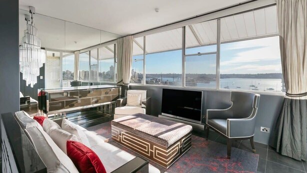 Ein modernes Wohnzimmer mit einer Couch, einem Couchtisch und einem Fernseher. Ein großes Fenster bietet Blick auf eine Stadtlandschaft und Wasser.