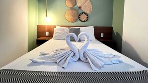 Una cama bien hecha con dos toallas dispuestas en forma de corazón, una lámpara colgante y una pared decorada con elementos tejidos.