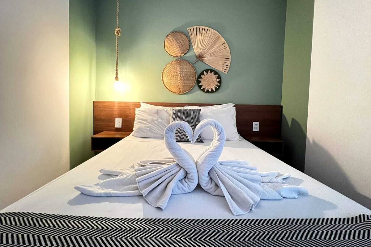 Una cama bien hecha con dos toallas dispuestas en forma de corazón, una lámpara colgante y una pared decorada con elementos tejidos.
