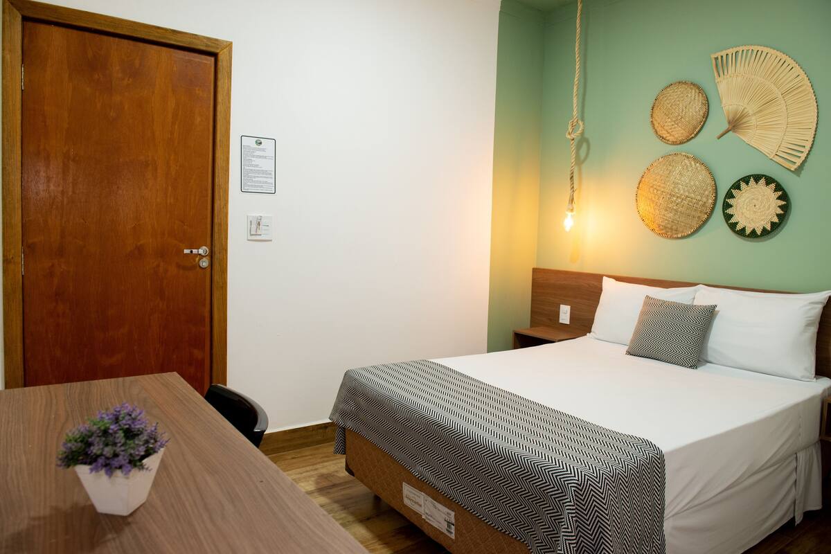 Un dormitorio con una cama, mesitas de noche, una puerta de madera, un ventilador de pared y una planta en maceta.