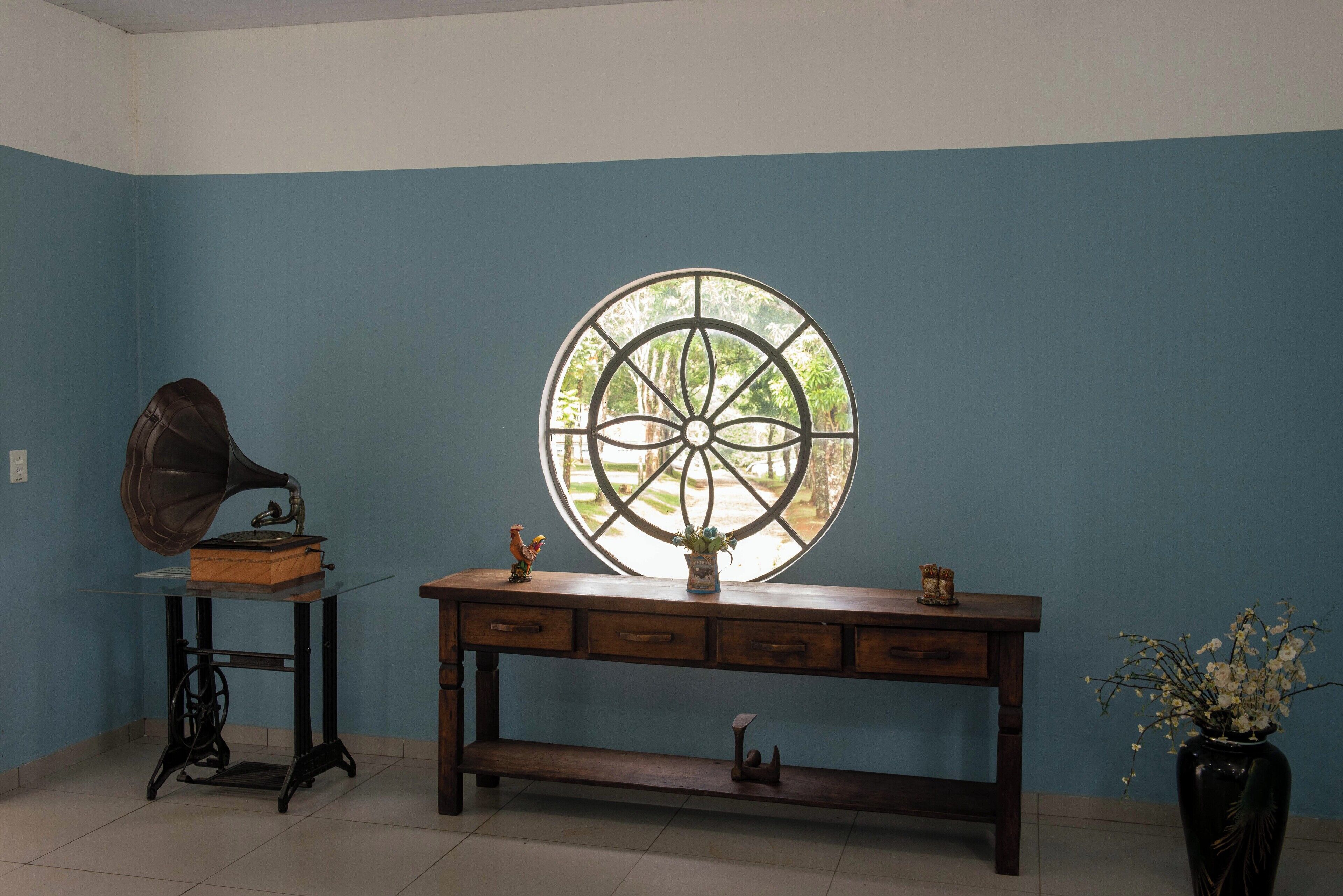 A room with a blue wall, a wooden console table with drawers, a vintage gramophone, a large circular window, and a vase with flowers.