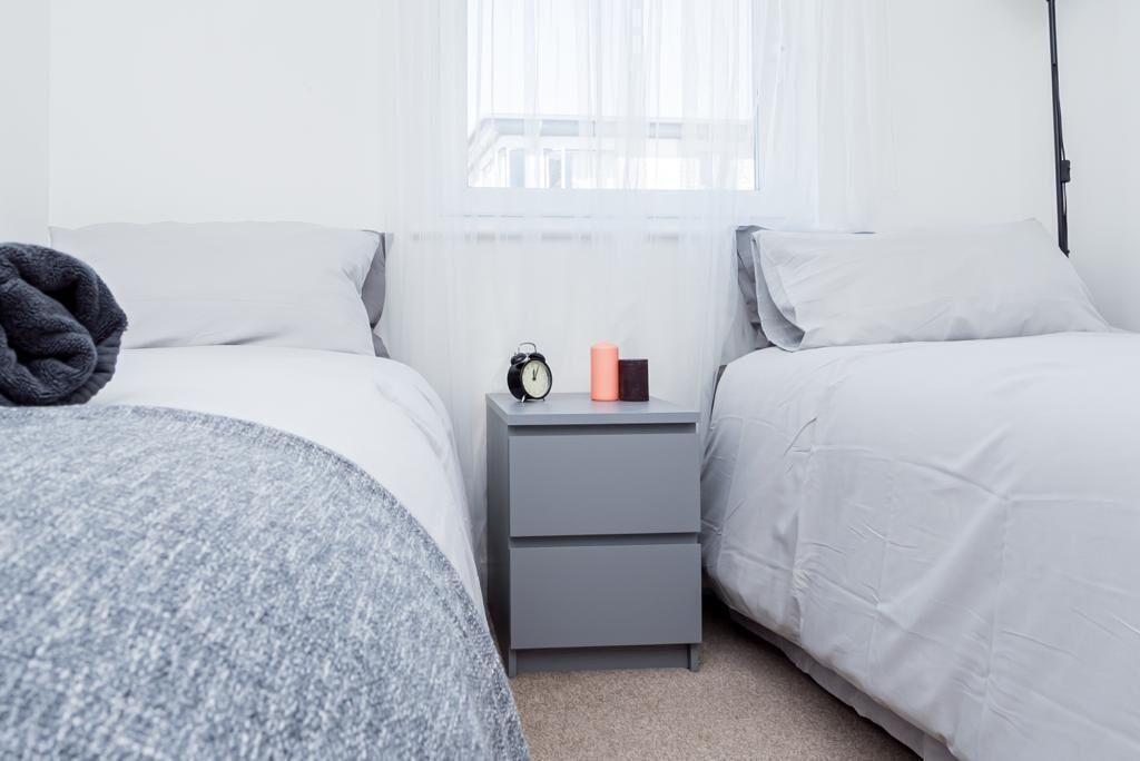 Un dormitorio con dos camas, una mesita de noche con un reloj y una vela, y cortinas translúcidas en la ventana.