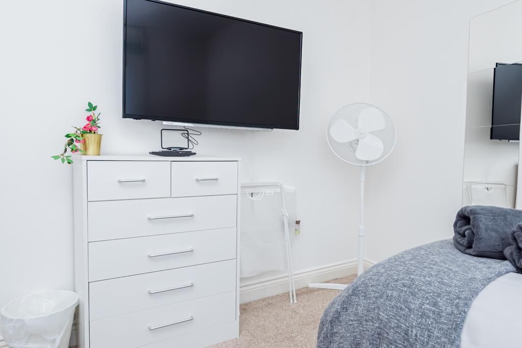 A modern bedroom with a flat-screen TV mounted on the wall, a white dresser with drawers, a bedside table, a grey bed with a grey blanket, and a white standing fan.