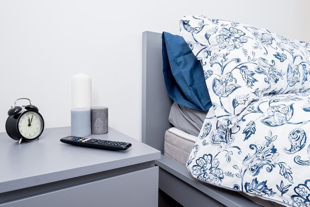 Un dormitorio ordenado con cama, mesita de noche, despertador y control remoto.