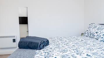Ein ordentlich bezogenes Bett mit einer blau-weißen blumigen Tagesdecke, einer aufgerollten grauen Decke und einer grauen Wolldecke auf einer hölzernen Bank.