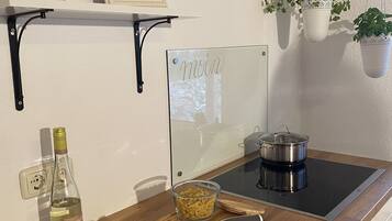 Eine Küche mit Holzarbeitsplatte, Schneidebrett, einer Weinflasche, einer Glasbowl mit Pasta, einem Messer und einem Topf auf einem Induktionskochfeld. Es gibt hängende Pflanzen und ein gerahmtes Foto an der Wand.