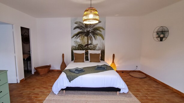 Een slaapkamer met een bed, een hanglamp, een wanddecoratie van palmbomen, een groene dressoir en een mand.