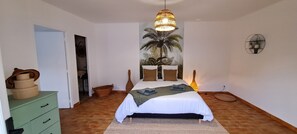 Un dormitorio con una cama, una lámpara colgante, un cuadro de palmeras, un aparador verde y una cesta.