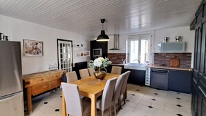 Een moderne keuken met een roestvrijstalen koelkast, een houten eettafel en stoelen. Er is een houten bankje met een metalen mand, een vaas met bloemen en een hanglamp.