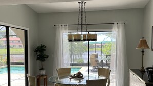 A dining area with a glass table and chairs, a hanging light fixture, a potted plant, and a view of a swimming pool. - Waterfront Spanish Villa with private pool and lake views - 5 mins from beach! (Bonita Springs)