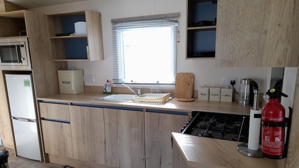 Cucina compatta con mobili in legno, forno a microonde integrato, lavandino e piano cottura a gas. Sono presenti ripiani portaoggetti e una finestra sopra il lavandino.