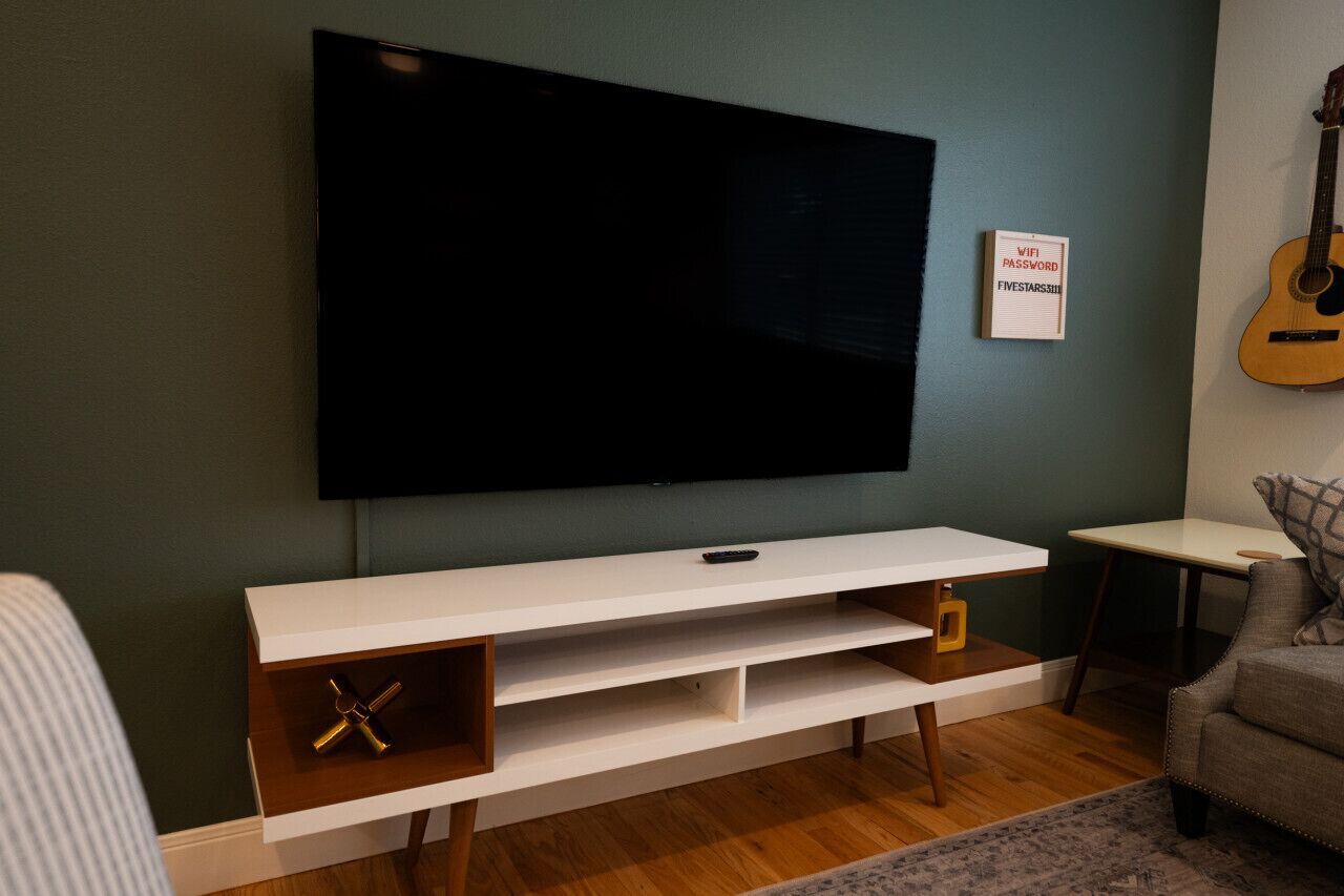 Een moderne woonkamer met een flatscreen televisie aan de muur, een witte entertainmentkast met planken, een gitaar aan de wand en een kleine tafel met een kussen.