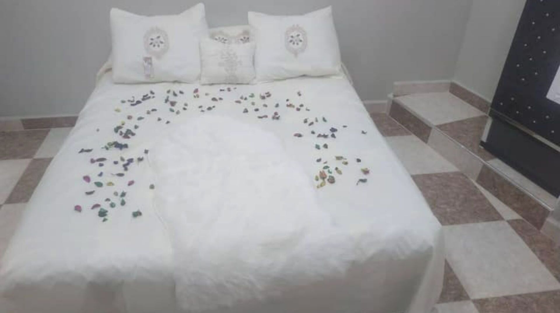 Una cama con cubrecama y almohadas blancas, suelo de baldosas y un mueble oscuro a la derecha.