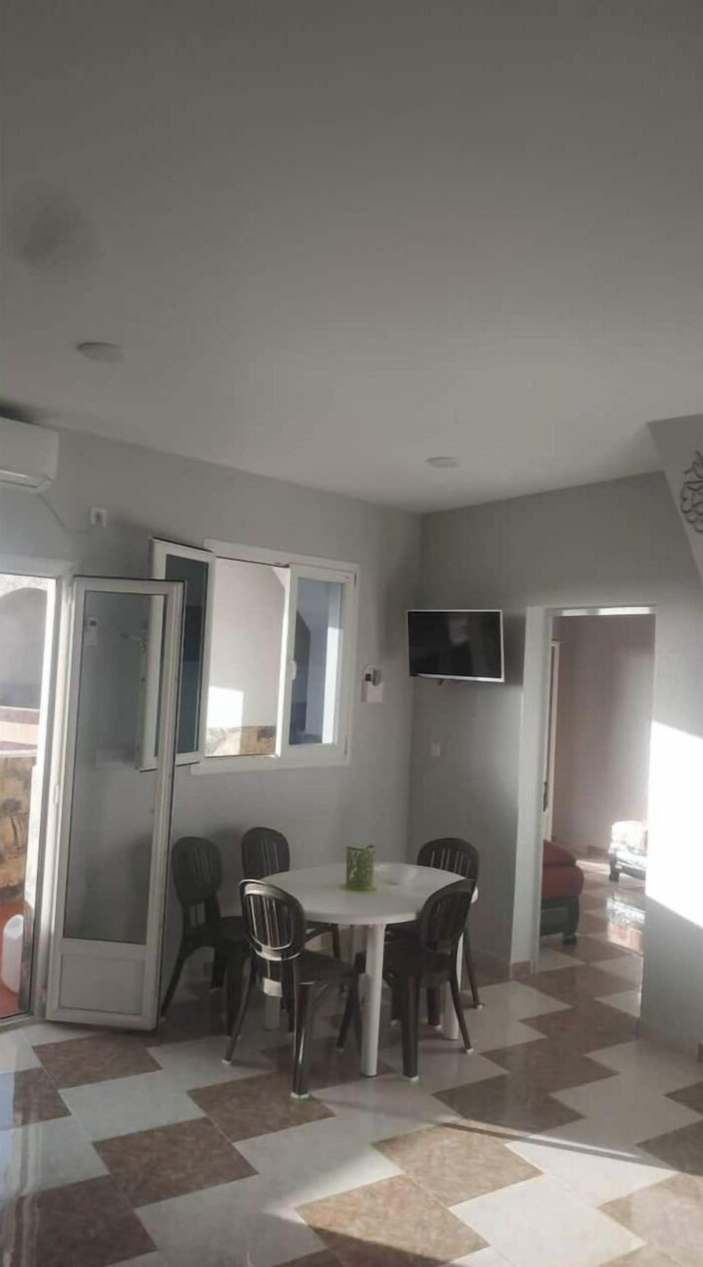 A dining area with a round table and four chairs, a television mounted on the wall, and a checkered floor pattern.