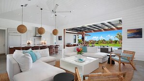 A spacious living area with a large window overlooking a lush garden, a ceiling fan, and a modern kitchen in the background.