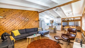 Een moderne woonruimte met een houten plafond, een bank, een salontafel, een leren fauteuil en een keuken met witte kasten en een magnetron.