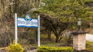 Bordje voor White Lake Homes met een logo, omringd door bomen en struiken.