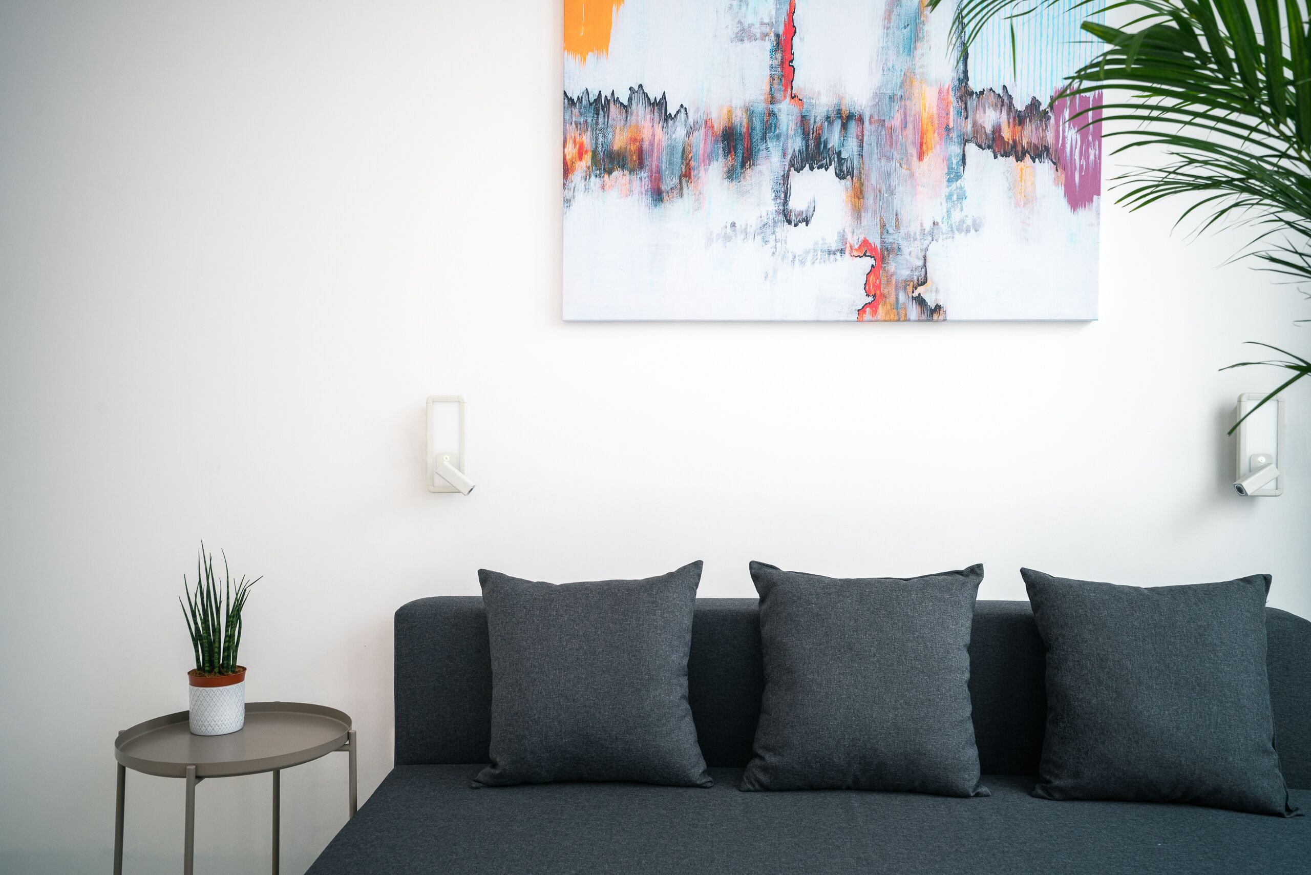 Ein modernes Wohnzimmer mit einem grauen Sofa, einem kleinen runden Tisch mit einer Pflanze und einem farbenfrohen, abstrakten Gemälde an der Wand.