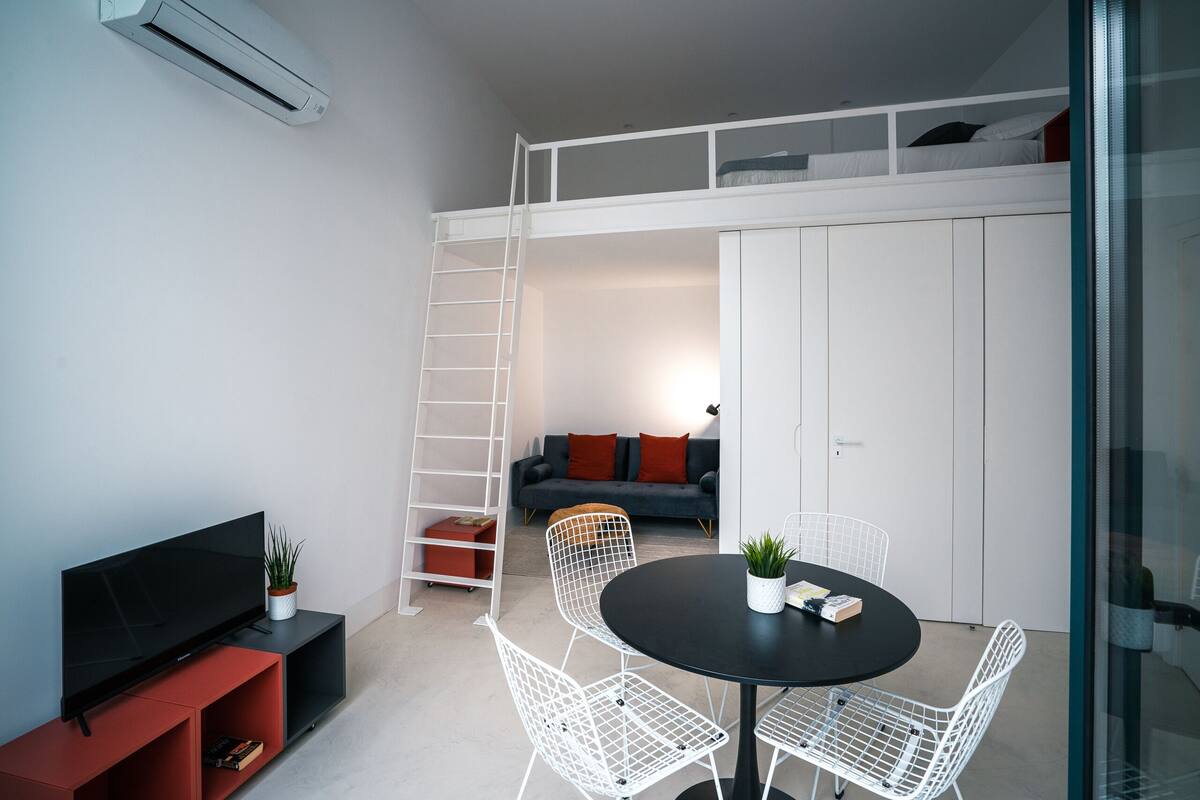 Ett modernt vardagsrum med en tv, vita stolsitsar av metalltråd, ett svart bord, en säng placerad på en loftnivå och en soffa med röda kuddar.