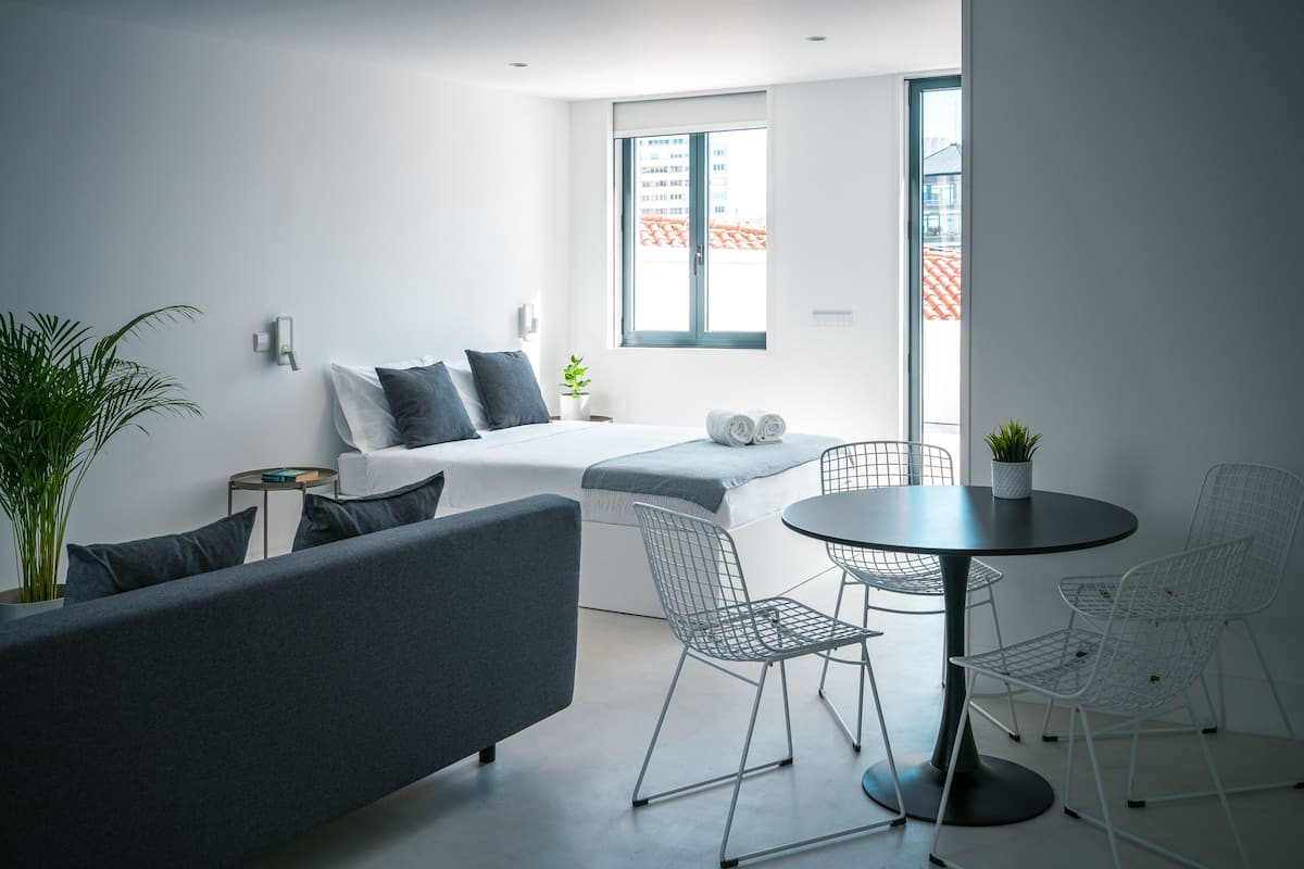 Ett modernt, minimalistiskt sovrum med en säng, en grå soffa och en liten matplats med metallstolar och ett bord.