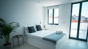 A modern bedroom with a bed, a small side table, a potted plant, and a window with a city view. - Portaria Housing (Aveiro)