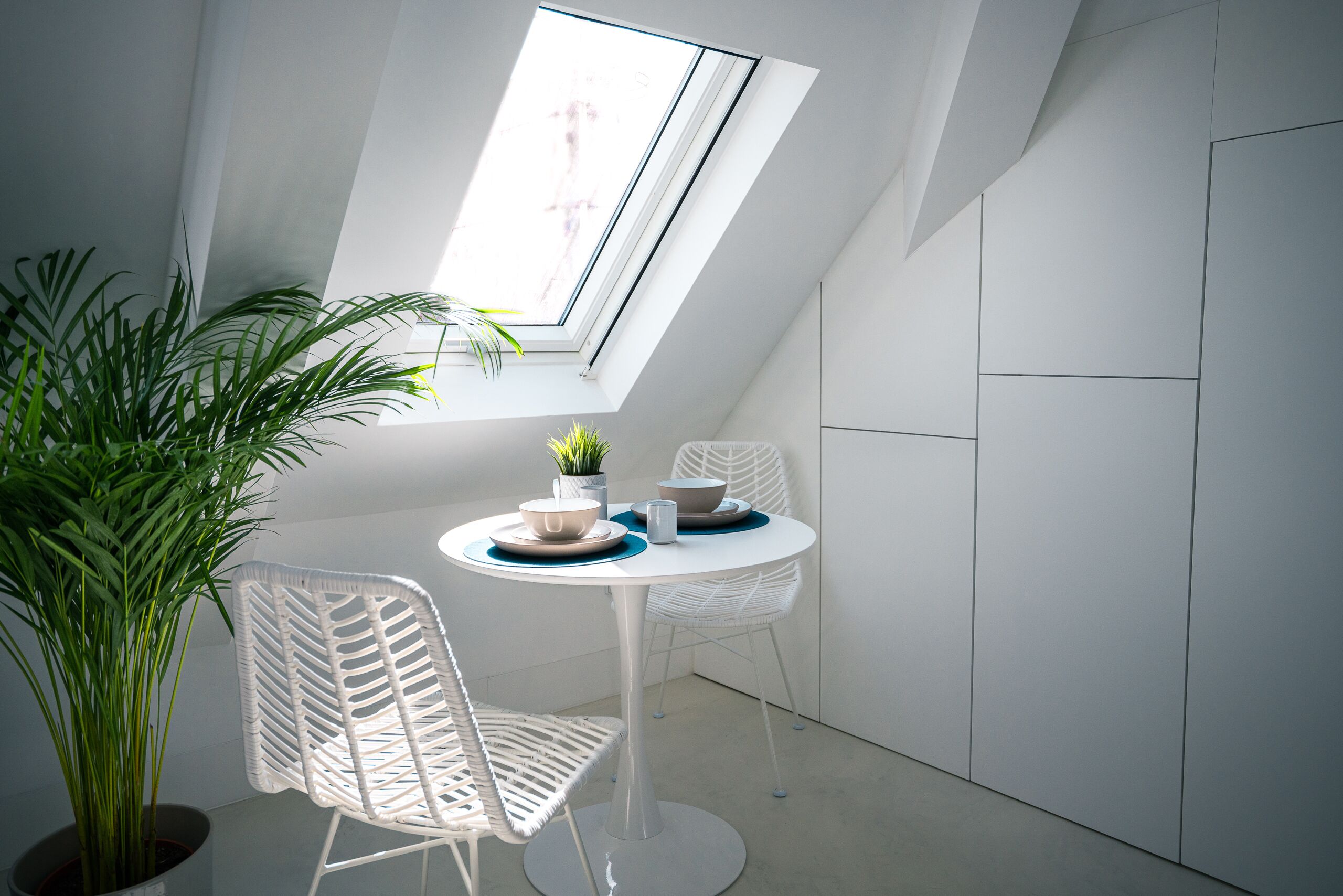 Une petite pièce moderne avec une lucarne, des chaises en osier blanches, une table ronde et une plante en pot.
