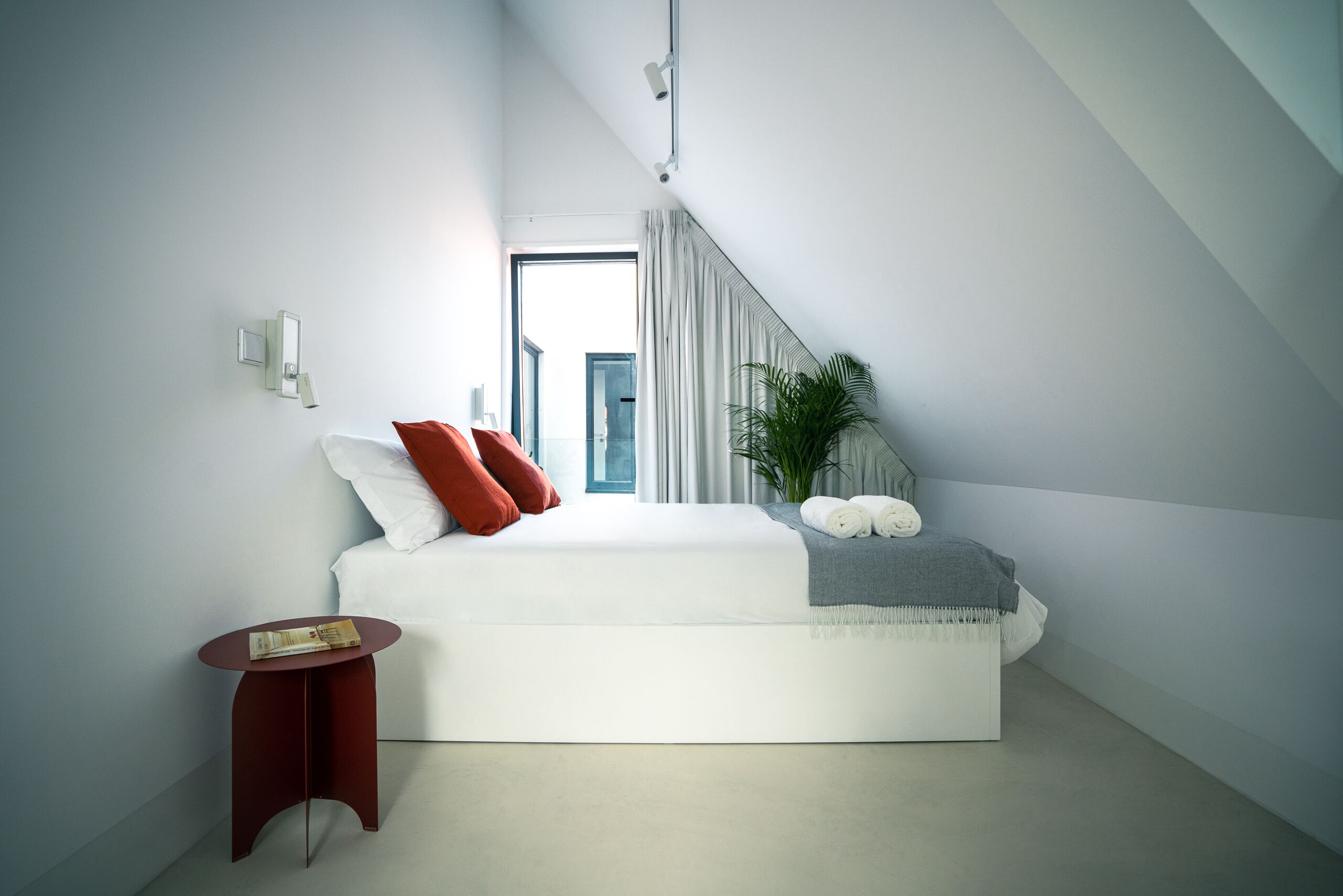 A modern bedroom with a large bed, a small side table, a potted plant, and a view of the city through a window.