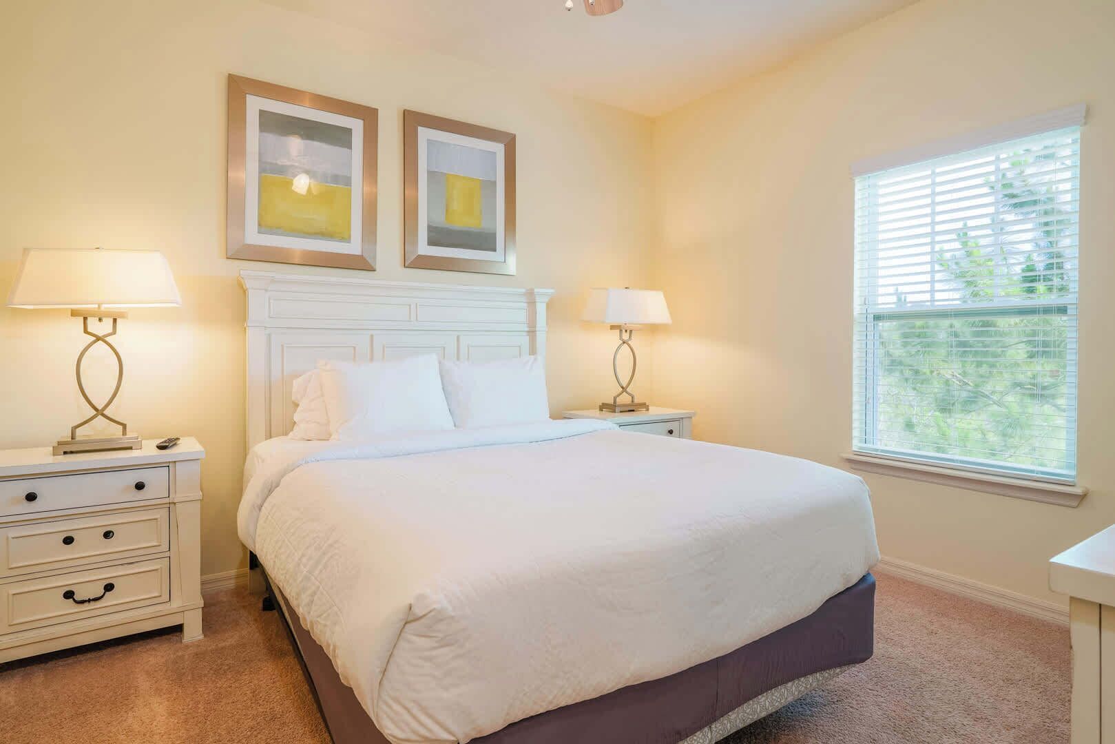 A bedroom with a bed, two bedside tables, a nightstand, a window with blinds, and two framed pictures on the wall.