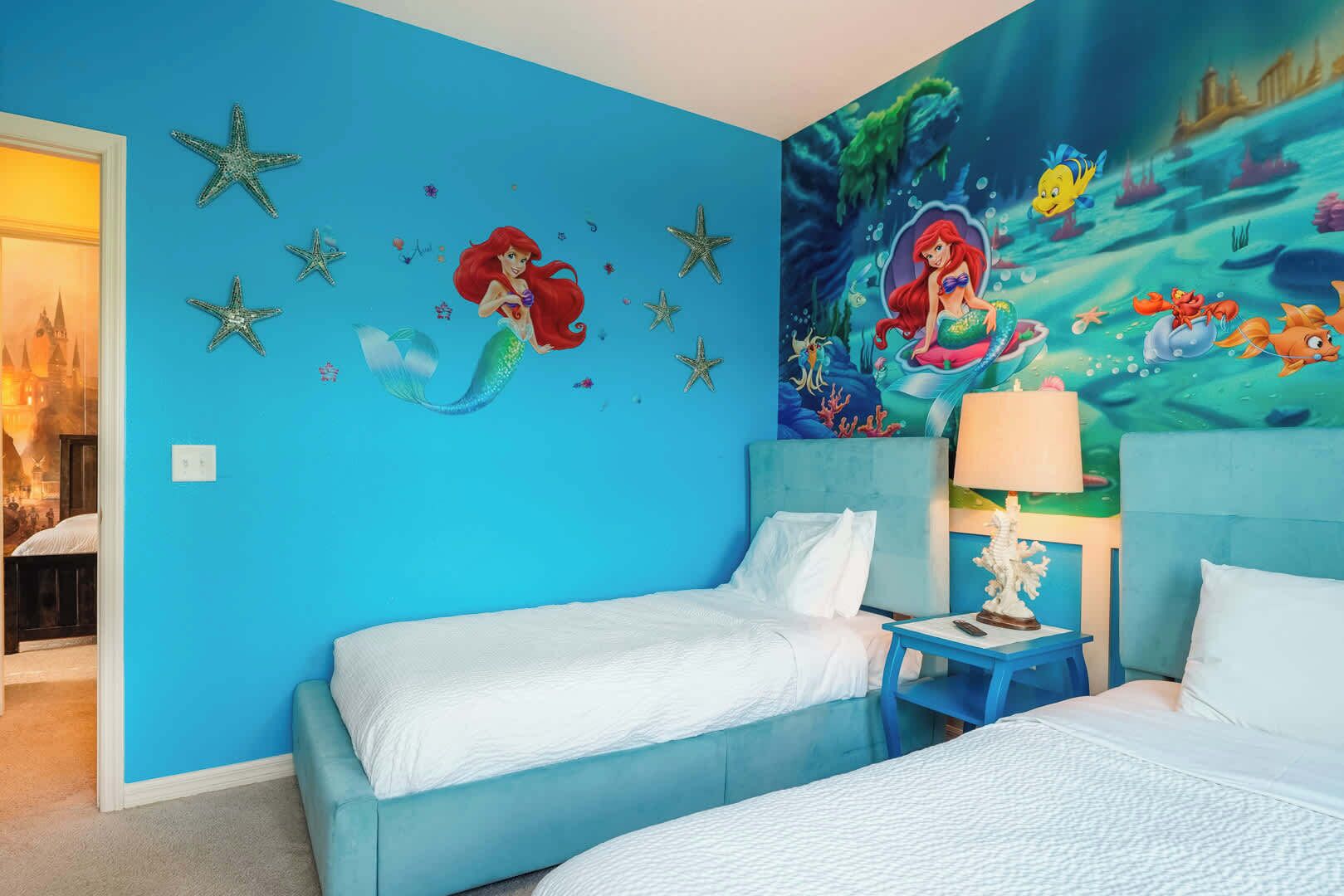 Una habitación con dos camas, un mural de sirenas, adornos de estrellas de mar y una lámpara en una mesita de noche.