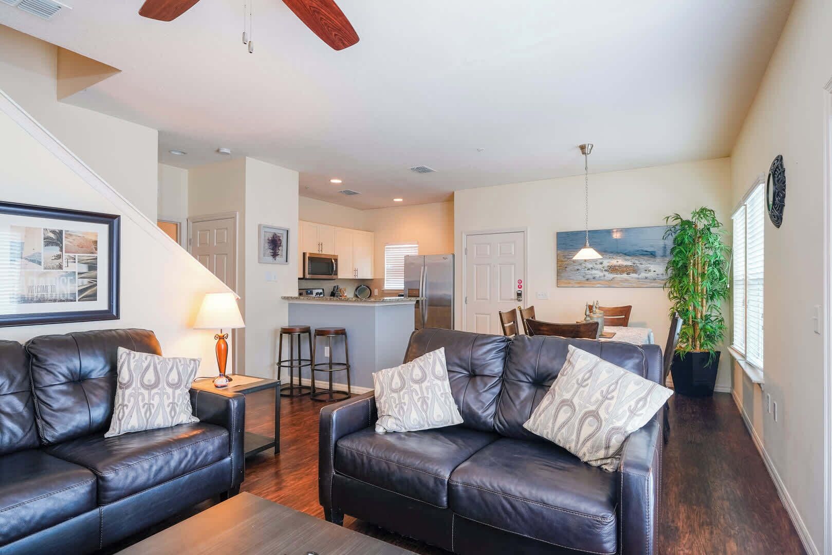 A living room with two leather sofas, a coffee table, and a lamp. There is a kitchen in the background with a refrigerator, microwave, and bar stools. A painting hangs on the wall, and a plant is in the corner.
